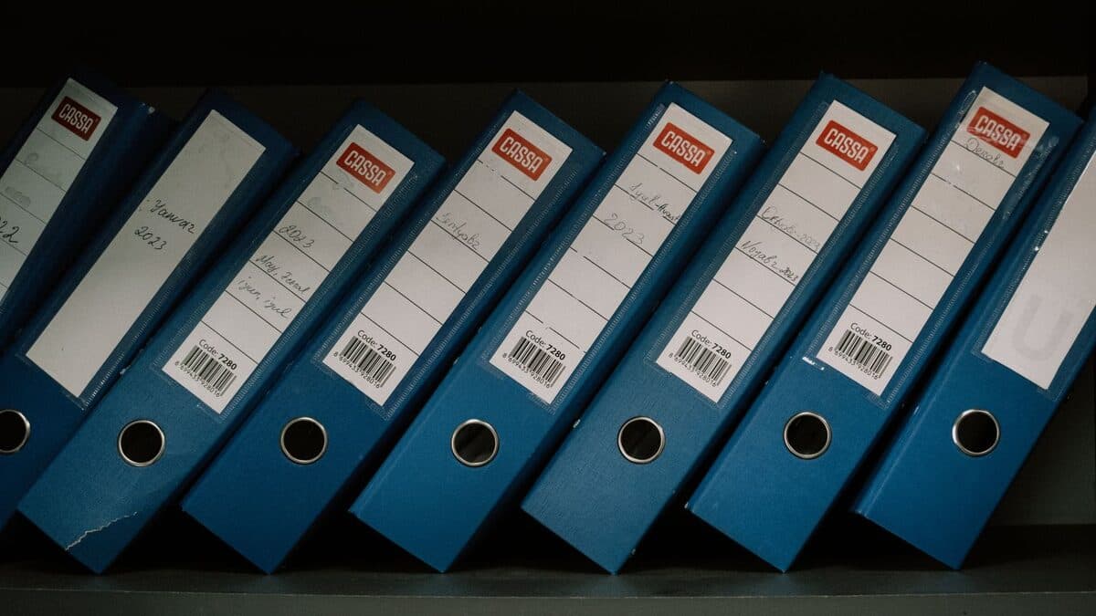 Classeurs d'archives d'entreprise étiquetés par dates et noms, rangés sur une étagère de bureau.